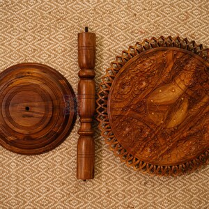 May include: A disassembled wooden side table with a round base, a central support, and a decorative top. The table components are a warm brown color and are arranged on a textured, neutral-toned surface.