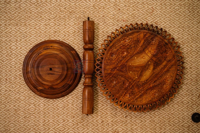 May include: A disassembled wooden side table with a round base, a central support, and a decorative top. The table components are a warm brown color and are arranged on a textured, neutral-toned surface.