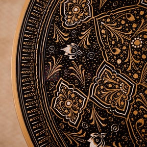 May include: Close-up of a decorative, round, black tray with intricate gold floral patterns. The tray's edge is also gold, with a detailed border design. The design includes white accents, creating a striking contrast.
