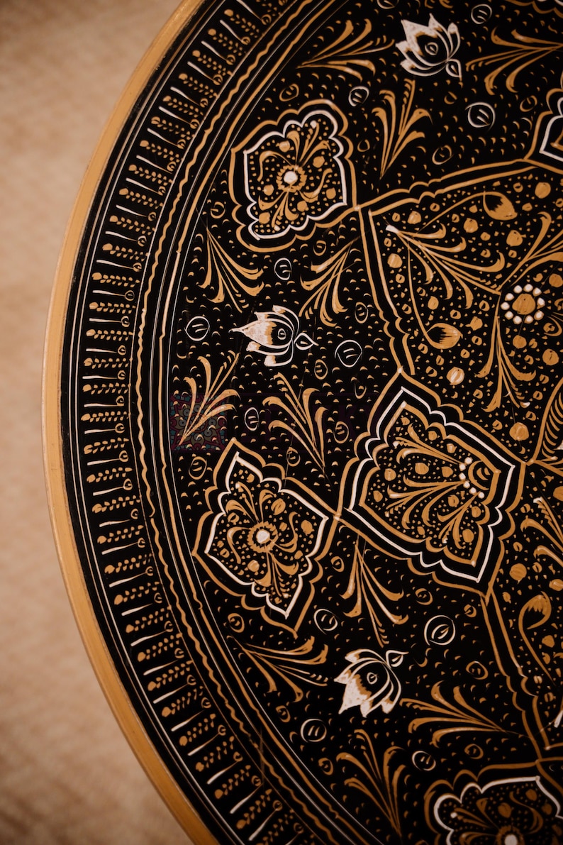 May include: Close-up of a decorative, round, black tray with intricate gold floral patterns. The tray's edge is also gold, with a detailed border design. The design includes white accents, creating a striking contrast.