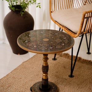 May include: A round, decorative side table with an ornate, patterned top. The table has a gold-colored rim and a dark brown center with floral and geometric designs. The table's base is gold and dark brown, and it stands on a woven rug.