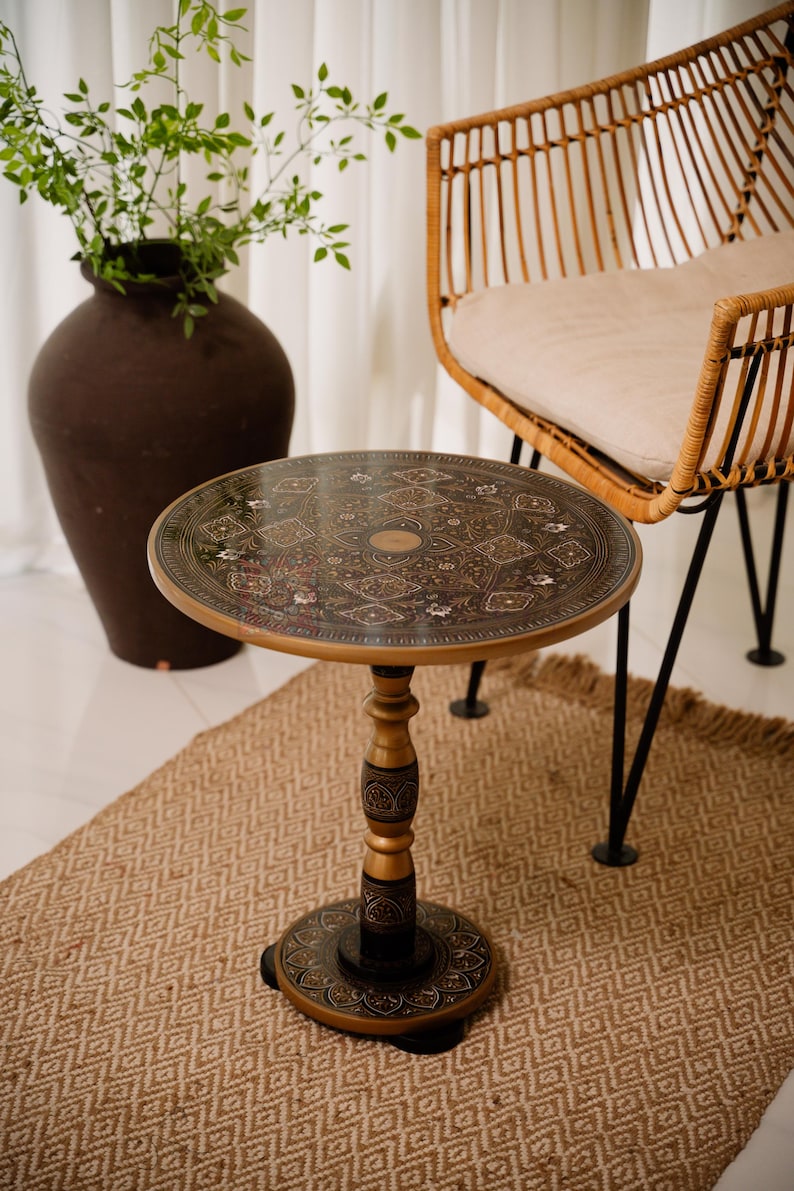 May include: A round, decorative side table with an ornate, patterned top. The table has a gold-colored rim and a dark brown center with floral and geometric designs. The table's base is gold and dark brown, and it stands on a woven rug.