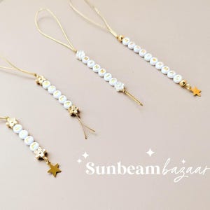 May include: Four gold-colored beaded charms with white beads spelling out a word, each with a gold star or flower charm at the end. The charms are connected to a thin gold string. The background is a light beige color.