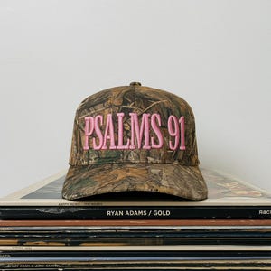 May include: A camouflage baseball cap with the text "PSALMS 91" embroidered in pink. The hat is sitting on top of a stack of vinyl records.