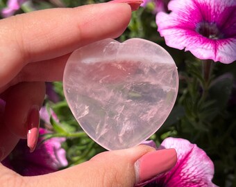 May include: A heart-shaped rose quartz crystal, polished smooth and translucent, with a light pink hue. The crystal is held in a hand against a backdrop of pink and white flowers.
