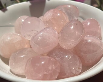 May include: A white bowl filled with tumbled rose quartz stones. The stones are smooth and have a light pink color.