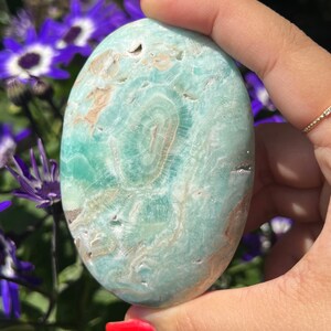 May include: A polished, oval-shaped gemstone in shades of teal and beige, held in a hand. The stone features a swirling pattern and is smooth to the touch. The background includes purple flowers.