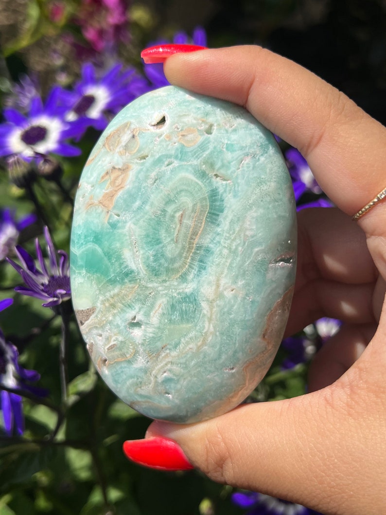 May include: A polished, oval-shaped gemstone in shades of teal and beige, held in a hand. The stone features a swirling pattern and is smooth to the touch. The background includes purple flowers.