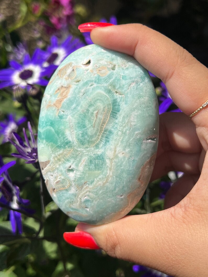 May include: A polished, oval-shaped stone with a light teal and tan color scheme. The stone has a smooth surface with natural patterns and is held in a hand with red nail polish. The background features purple flowers.