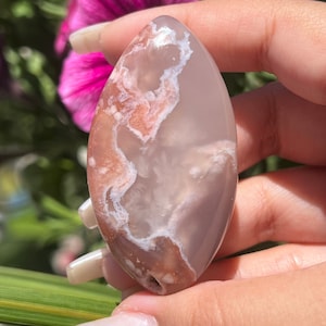 May include: A smooth, oval-shaped agate stone with a white and pink swirling pattern. The stone is translucent and has a light pink hue.