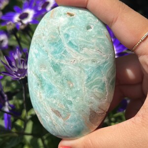 May include: A smooth, oval-shaped, light blue stone with white and brown veining. The stone is held in a hand with red nail polish, with purple flowers in the background. The stone's surface has a polished, glossy appearance.