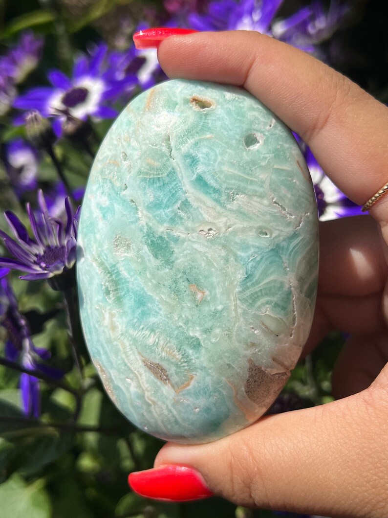 May include: A smooth, oval-shaped, light blue-green stone with a marbled appearance. The stone has a polished surface with natural variations in color and texture, and is held in a hand with red nail polish. The background features purple flowers.