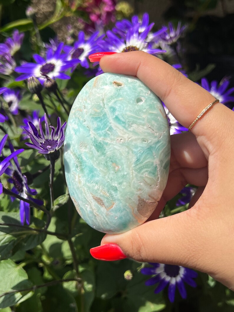 May include: A smooth, oval-shaped stone with a light blue and white marbled pattern. The stone is held in a hand with red nail polish, with purple and white flowers in the background.