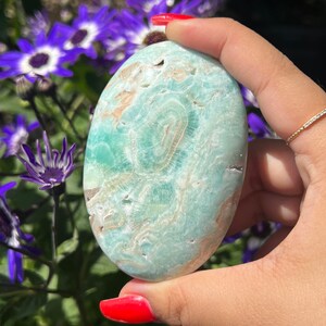 May include: A smooth, oval-shaped stone with a light teal and tan pattern. The stone is held in a hand with red nail polish, with purple and white flowers in the background. The stone's surface has a swirling, natural design.