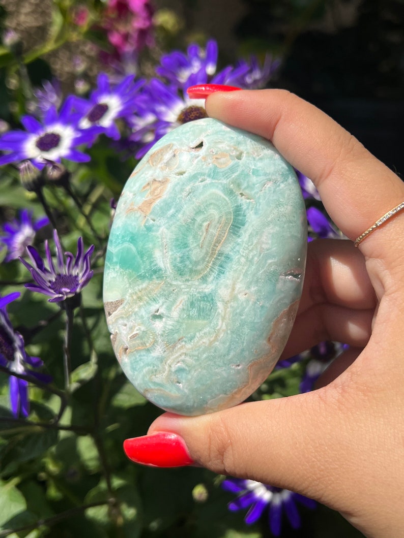 May include: A smooth, oval-shaped stone with a light teal and tan pattern. The stone is held in a hand with red nail polish, with purple and white flowers in the background. The stone's surface has a swirling, natural design.
