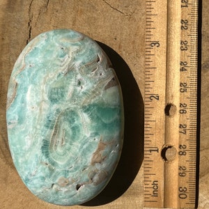 May include: An oval-shaped, polished stone with a swirling pattern of light green, white, and brown hues. The stone is next to a wooden ruler, showing measurements in inches.