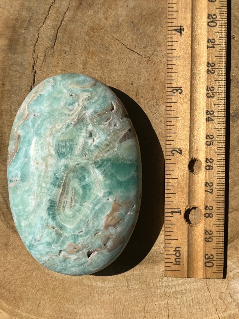 May include: An oval-shaped, polished stone with a swirling pattern of light green, white, and brown hues. The stone is next to a wooden ruler, showing measurements in inches.