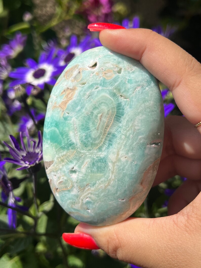 May include: A smooth, oval-shaped stone with a light blue and tan pattern. The stone is held in a hand with red nail polish, with purple flowers in the background. The stone's surface has a swirling, natural design.