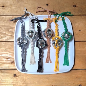 May include: A collection of seven disco ball ornaments, each with a unique macrame hanger in various colors: gray, white, black, tan, yellow, and green. The ornaments are displayed on a white rectangular plate against a wooden background.