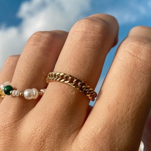Peut inclure: Deux bagues en or sur une main. L'une est une simple chaîne à maillons. L'autre est une chaîne délicate avec une pierre verte et des perles blanches.