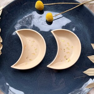 Può includere: Due ciotole a forma di mezzaluna in ceramica beige con dettagli in foglia d'oro. Su un piatto blu scuro, con elementi floreali secchi e una foglia di palma. Design minimalista e decorativo, adatte per piccoli snack.