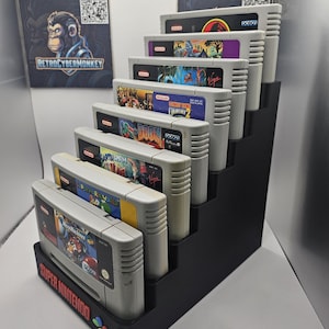 Op de afbeelding: Een zwarte, getrapte plastic displaystandaard met zeven grijze Super Nintendo-spelcartridges. De voorkant van de standaard is voorzien van de rode tekst "SUPER NINTENDO" en een klein, meerkleurig icoon van een controllerknop. De cartridges zijn in een trapvorm gerangschikt.