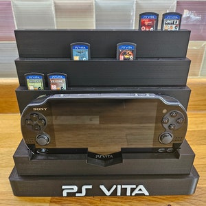 Peut inclure: Un support en plastique noir pour une console de jeu portable PlayStation Vita. Le support a un emplacement pour la console et une étagère pour ranger les cartouches de jeu. Le support porte l'inscription "PS VITA" sur le devant.