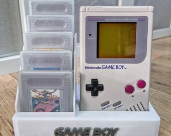 Puede incluir: Un soporte de plástico blanco con una consola Game Boy gris y cinco cartuchos de juego. El soporte tiene las palabras "GAME BOY" impresas en la parte delantera.