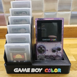 Może przedstawiać: Czarny stojak wystawowy Game Boy Color z przezroczystą fioletową konsolą Game Boy Color i kilkoma kartridżami do gier. Stojak ma białe słowa "GAME BOY COLOR" z kolorowymi literami. Widoczny jest kartridż do gry The Lion King.