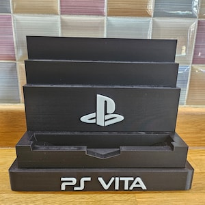 Peut inclure: Un support en plastique noir avec un logo PlayStation Vita et les mots "PS VITA" imprimés sur le devant. Le support a une fente pour une console de jeu portable.