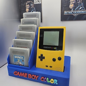 Può includere: Una console portatile Game Boy Color giallo brillante è posizionata in un espositore in plastica blu. Il supporto presenta una rastrelliera a gradini che contiene sei cartucce di gioco grigie e la scritta GAME BOY COLOR in rilievo.