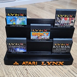 Könnte beinhalten: Ein schwarzer Atari Lynx-Spielkartuschenständer mit fünf Spielkartuschen. Die Kartuschen sind für die Spiele Awesome Golf, Paperboy, Tournament Cyberball, California Games und Batman Returns.