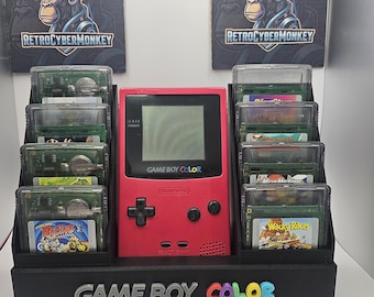 May include: A berry-colored Game Boy Color handheld console centered in a black display stand. The stand features the text "GAME BOY COLOR" in raised letters and holds eight game cartridges in clear plastic cases. Two "RETROCYBERMONKEY" signs with QR codes are positioned behind the console.