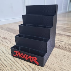 Può includere: Un supporto in plastica nera con cinque fessure per riporre oggetti. Il supporto ha la scritta "JAGUAR" stampata in rosso sul davanti.