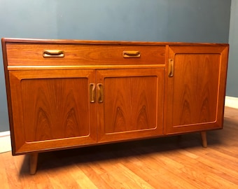 May include: A mid-century modern teak sideboard with three doors and one drawer. The cabinet has brass handles and a wood grain finish.