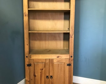 May include: A tall wooden bookcase with three shelves and two doors. The bookcase is made of light brown wood and has black metal hinges and handles.