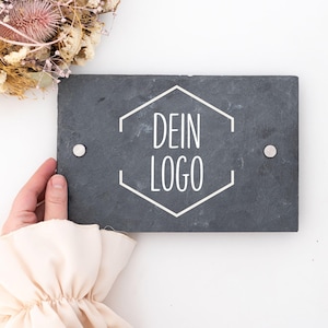 Könnte beinhalten: Ein schwarzes Schiefer-Schild mit abgerundeten Ecken und einem weißen sechseckigen Rahmen. Der Text "DEIN LOGO" ist in weiß innerhalb des Rahmens geschrieben.