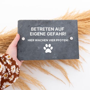 Könnte beinhalten: Schwarzes Schiefer-Schild mit weißem Text "Betreten auf eigene Gefahr! Hier Wachen Vier Pfoten!" und einem weißen Pfotenabdruck unter dem Text. Das Schild wird von einer Hand gehalten.