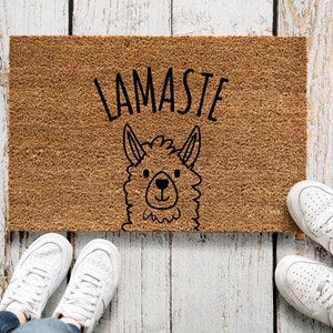 Op de afbeelding: Een bruine deurmat met de tekst "LAMASTE" en een zwart-witte illustratie van een lama-gezicht.