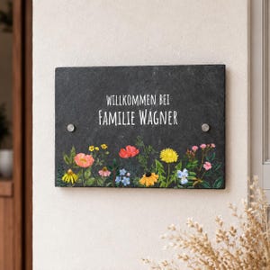 Op de afbeelding: Een rechthoekig zwart leistenen bord met de witte tekst WILLKOMMEN BEI FAMILIE WAGNER. Het onderste gedeelte van het bord is versierd met een kleurrijke illustratie van diverse wilde bloemen, waaronder gele, roze, rode en blauwe bloesems.