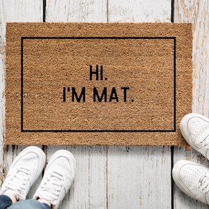 Könnte beinhalten: Eine braune Fußmatte mit dem Text "Hi. I'm Mat." in schwarzen Buchstaben. Zwei Paar weiße Turnschuhe stehen auf dem Holzboden.