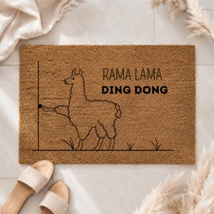 Könnte beinhalten: Eine rechteckige braune Kokosfasermatte mit einer schwarzen Strichzeichnung eines Lamas vor einem Türrahmen. Das Lama streckt die Pfote aus, um eine Türklingel zu drücken. Der Text RAMA LAMA DING DONG ist in schwarzen Buchstaben rechts aufgedruckt.