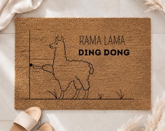 Op de afbeelding: Een rechthoekige bruine kokosmat met een zwarte lijntekening van een lama bij een deurkozijn. De lama reikt uit om op een deurbel te drukken. De tekst RAMA LAMA DING DONG staat in zwarte letters aan de rechterkant van de deurmat gedrukt.