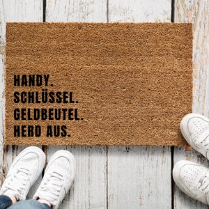 Pode incluir: Um tapete de coco castanho com o texto "HANDY. SCHLÜSSEL. GELDBEUTEL. HERD AUS." impresso em preto.