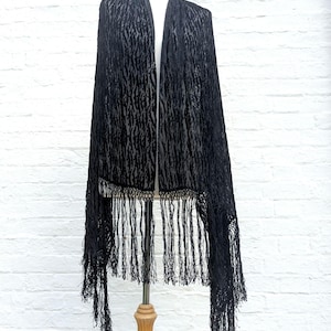 May include: A black shawl featuring a textured, velvet-like animal print pattern throughout the fabric. The garment is finished with long, thin black fringe detailing along the bottom edges, creating a draped appearance on a display mannequin.