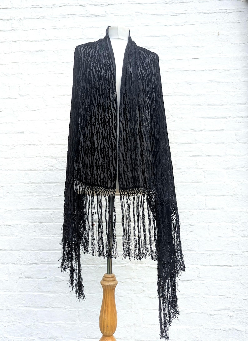 May include: A black shawl featuring a textured, velvet-like animal print pattern throughout the fabric. The garment is finished with long, thin black fringe detailing along the bottom edges, creating a draped appearance on a display mannequin.