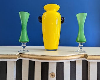 May include: Three decorative glass vases. A tall, yellow vase with dark blue handles is flanked by two green hourglass-shaped vases. The vases are displayed on a white and black striped cabinet against a blue wall.