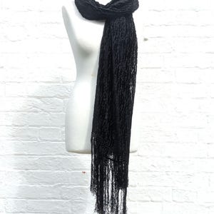 May include: A long black scarf featuring a textured, crinkled fabric surface and delicate fringe detailing along the bottom edge. The scarf is draped around the neck of a white mannequin, showcasing its length and intricate, semi-sheer pattern.