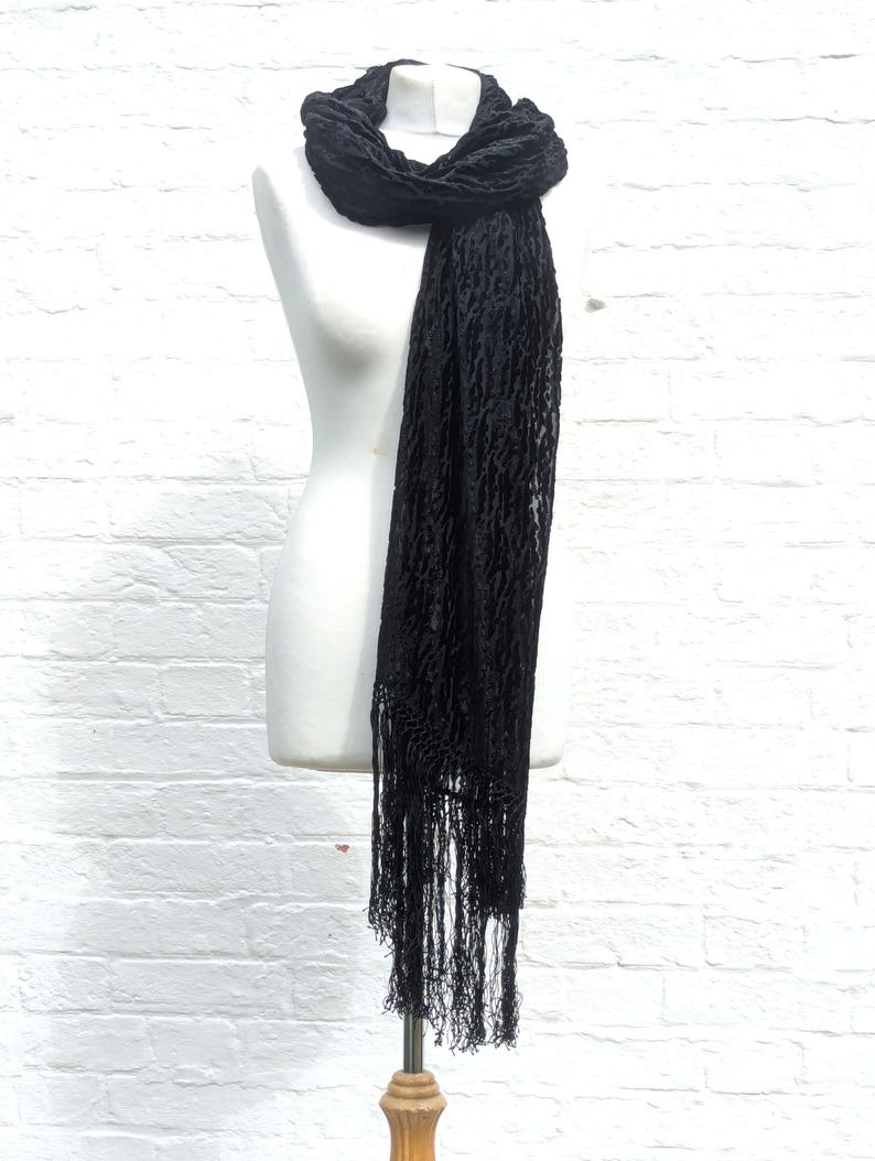 May include: A long black scarf featuring a textured, crinkled fabric surface and delicate fringe detailing along the bottom edge. The scarf is draped around the neck of a white mannequin, showcasing its length and intricate, semi-sheer pattern.
