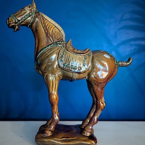 Puede incluir: Figura de caballo de cerámica en tonos marrón, beige y verde. El caballo está posicionado con la cabeza alzada y tiene una silla de montar. La figura está sobre una base rectangular y tiene un acabado brillante. El fondo es azul liso.